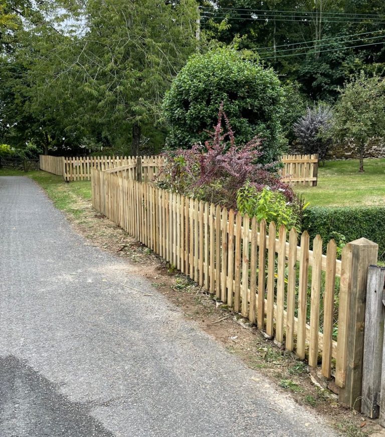Fencing Services Exterior photo of a light coloured wooden fence with well established plants and trees in the background.